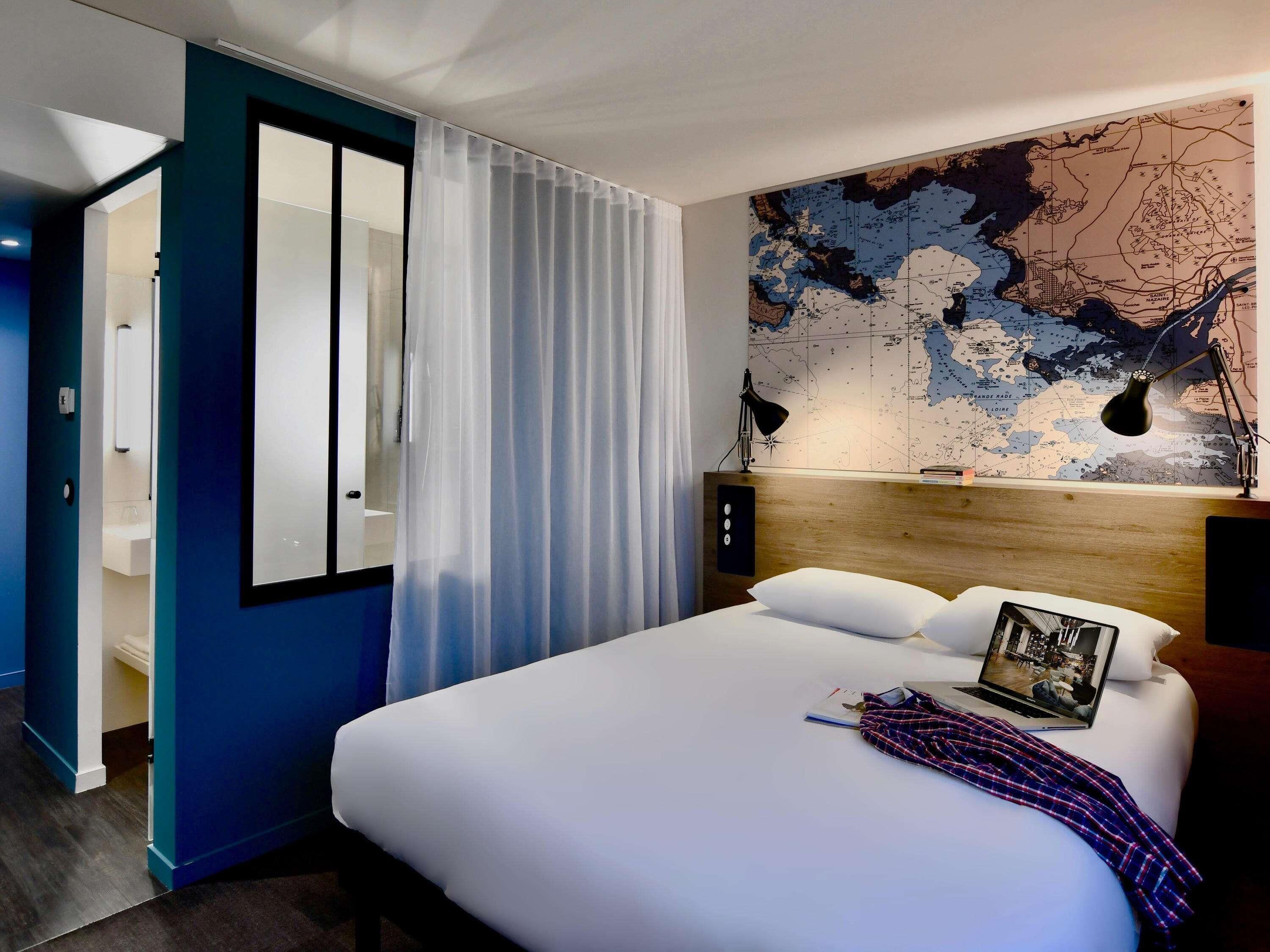 A hotel room with a bed, a map on the wall, a desk with a lamp, and a door leading to a bathroom.