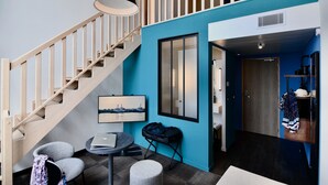 A modern room with a staircase, a desk with a laptop, a chair, a hanging light fixture, and a large window with blinds.