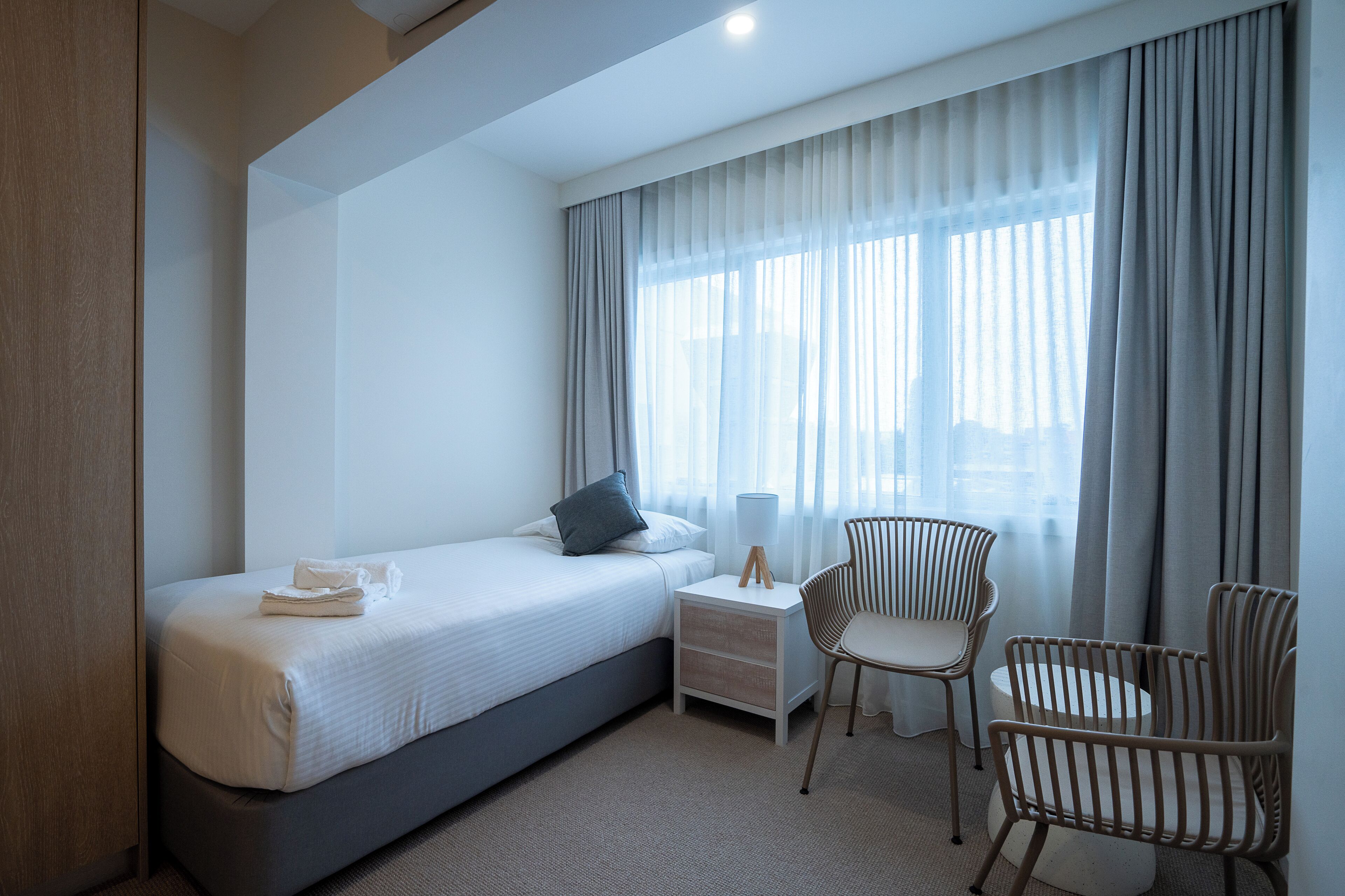 A hotel room with a bed, bedside table, lamp, two chairs, and a window with curtains.