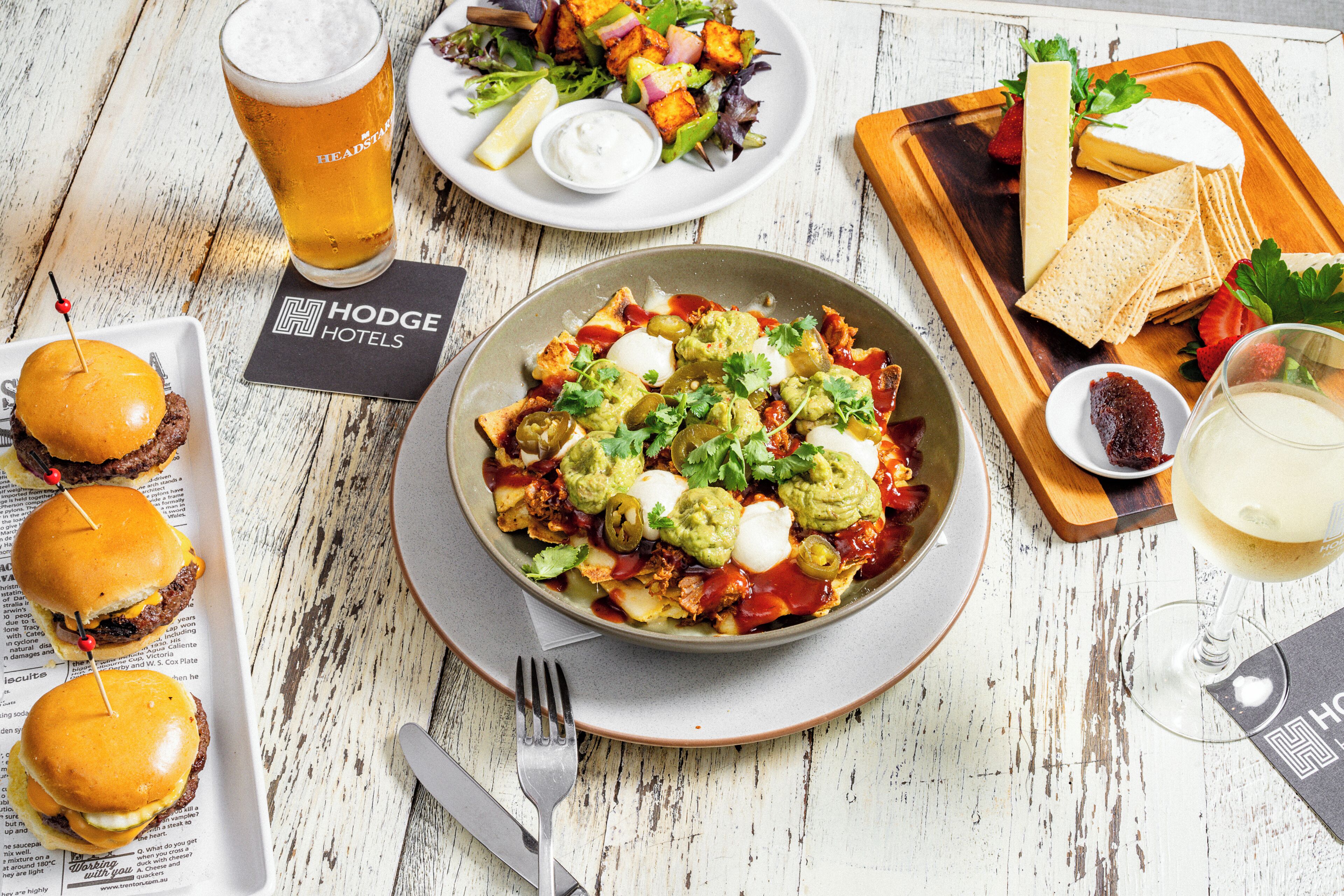 Une table à manger est dressée avec divers plats, notamment des hamburgers, une salade et un bol de guacamole aux œufs. On y trouve également des verres de bière et un verre à vin.