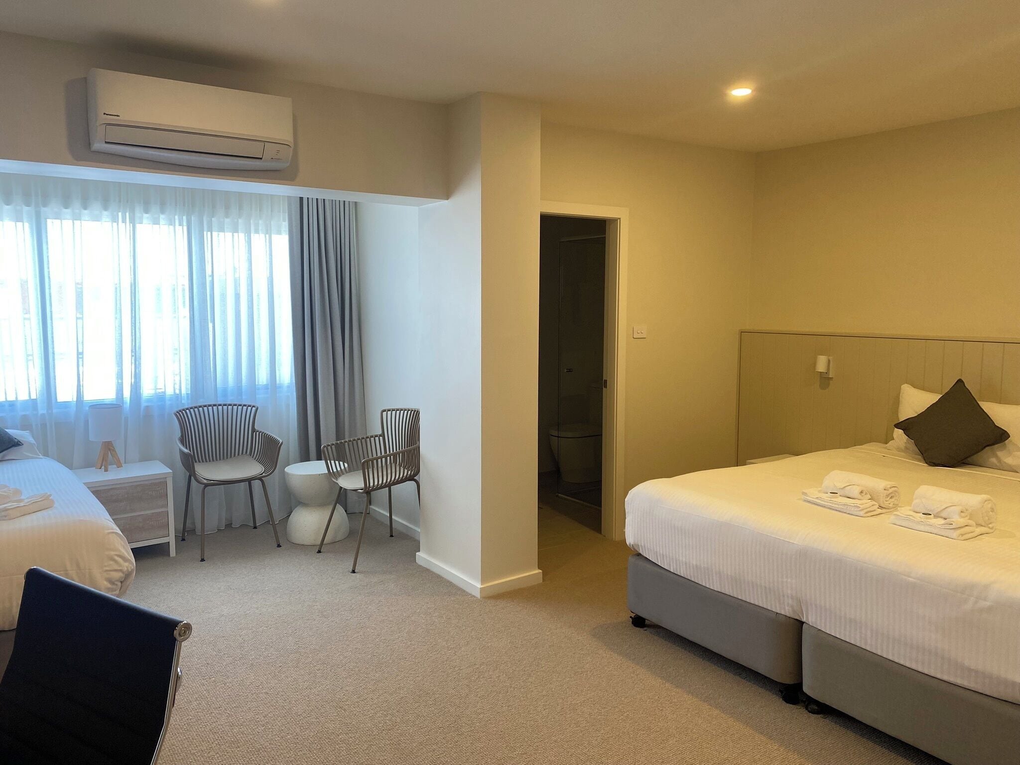 A hotel room with a bed, two chairs, a small table, an air conditioning unit, and a bathroom door.