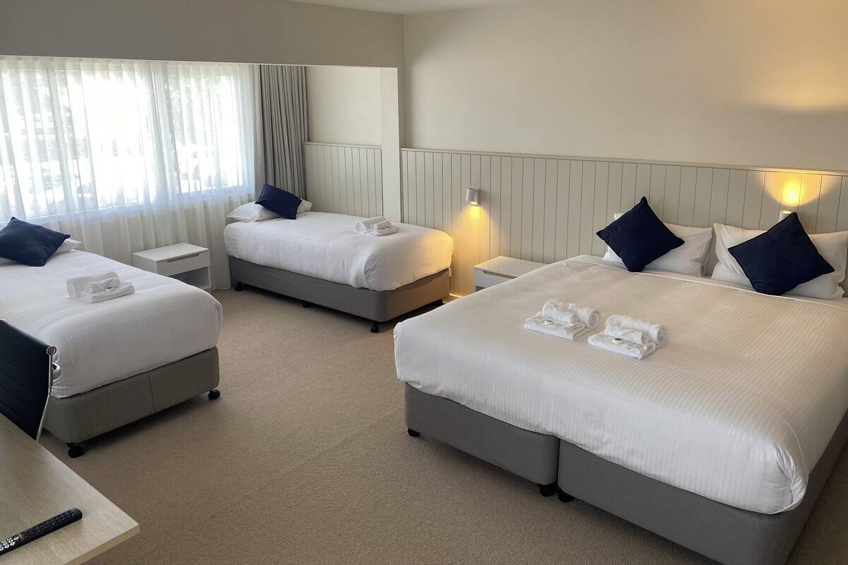 Ein Hotelzimmer mit zwei Betten, jeweils mit weißen Laken und dunklen Kissen, einem Nachttisch mit Fernbedienung und einem Fenster mit durchsichtigen Vorhängen.