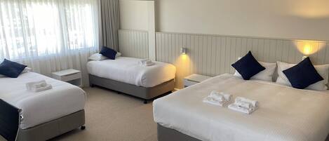 A hotel room with two beds, each with white linens and dark pillows, a bedside table with a remote control, and a window with sheer curtains.