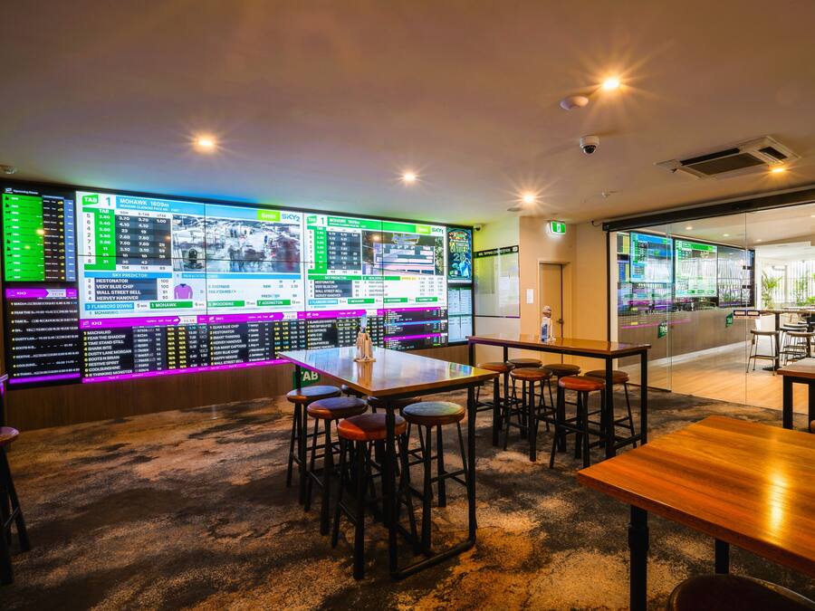 Zona de bar moderna con varias pantallas mostrando información sobre apuestas deportivas, taburetes de bar y una mesa de madera.
