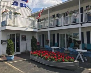 Hotel de dois andares com varanda no andar superior, área de estar externa e um canteiro de flores no centro.