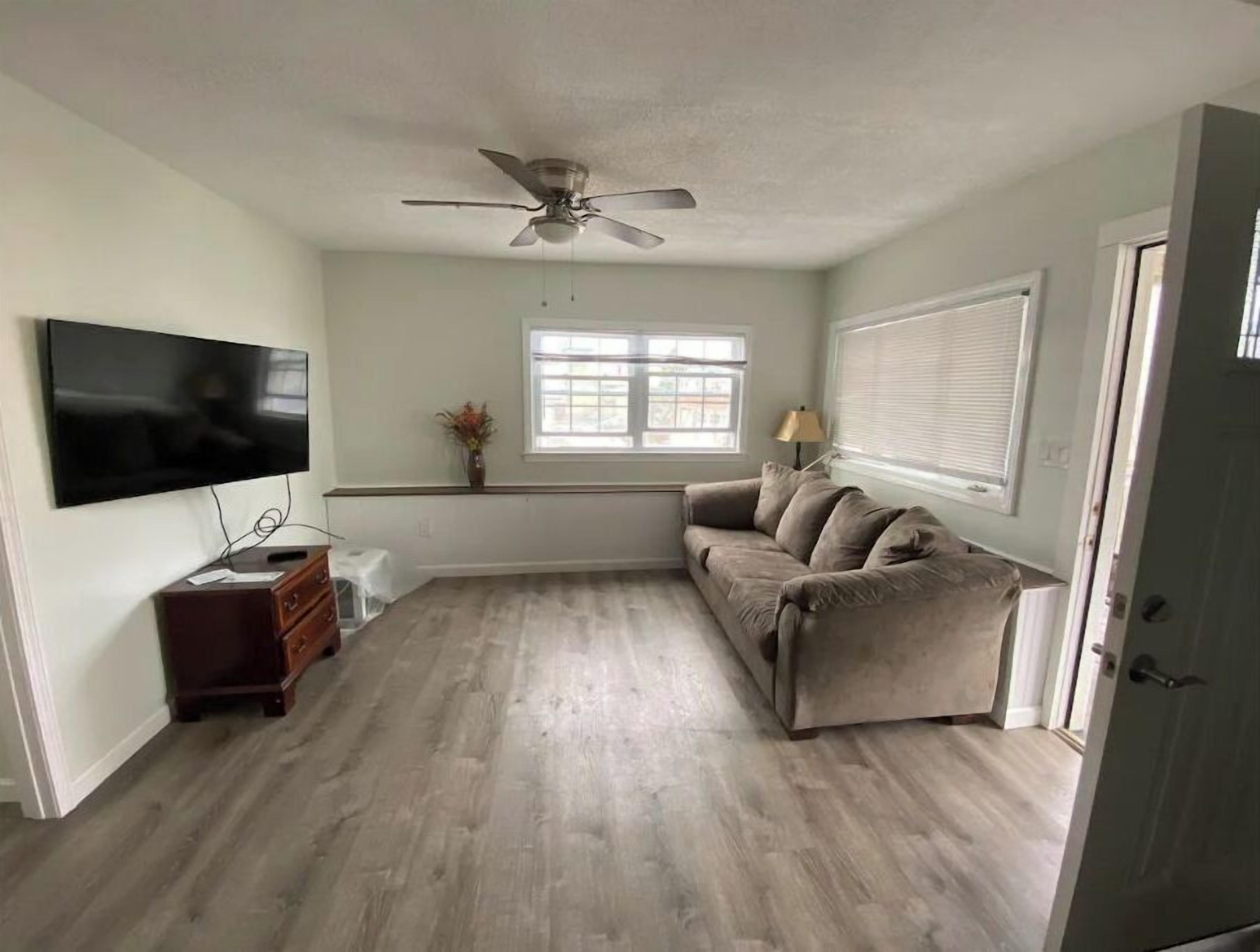 Un salon équipé d’un téléviseur à écran plat, d’un ventilateur de plafond, d’un canapé, d’une petite table et d’une fenêtre avec des stores.