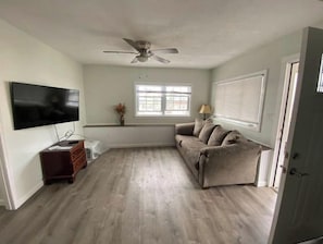 Uma sala de estar com televisão de tela plana, ventilador de teto, sofá, mesa pequena e janela com persianas.