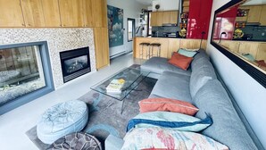 A modern living room with a fireplace, a sofa, ottomans, and a kitchen area visible through an open door.