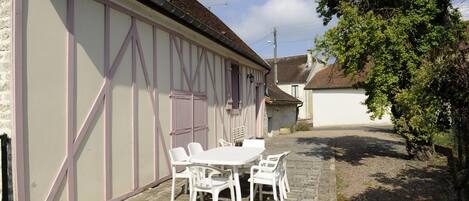 A building with a white exterior and purple accents, a paved courtyard with a table and chairs, and a large tree to the right.