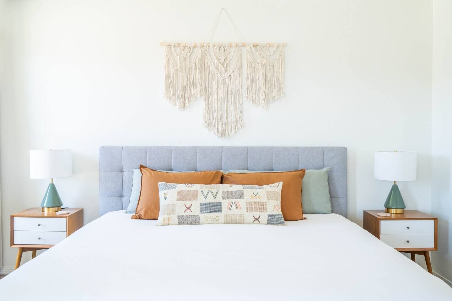Une chambre à coucher avec une tête de lit rembourrée de couleur grise, un couvre-lit blanc et deux tables de chevet avec des lampes.