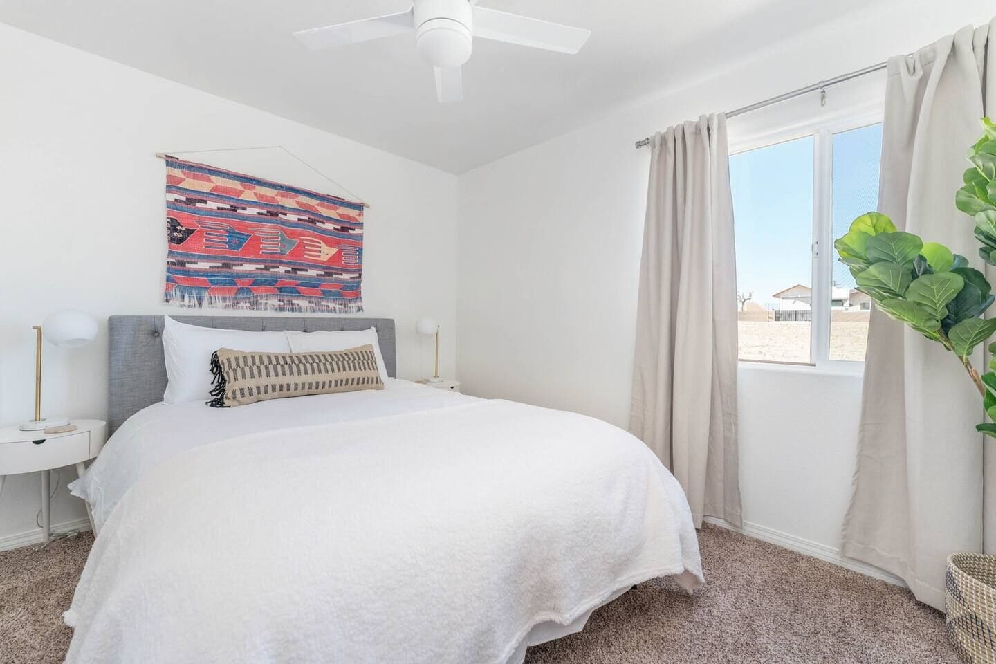 A bedroom with a bed, a ceiling fan, a window with curtains, a wall hanging, and a plant.