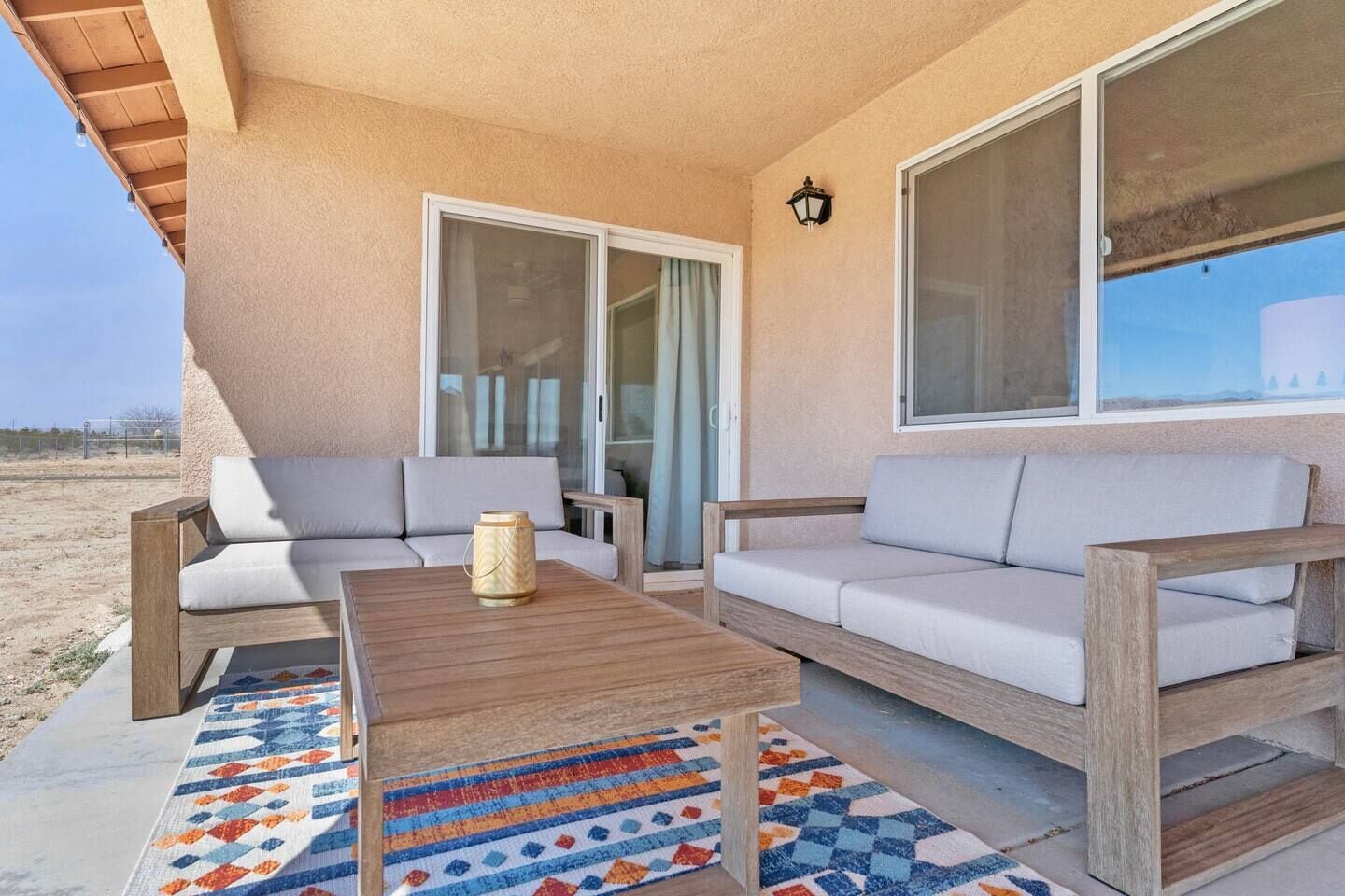Uma área de estar ao ar livre coberta, com dois sofás e uma mesa de madeira, um tapete colorido e um candeeiro na parede.
