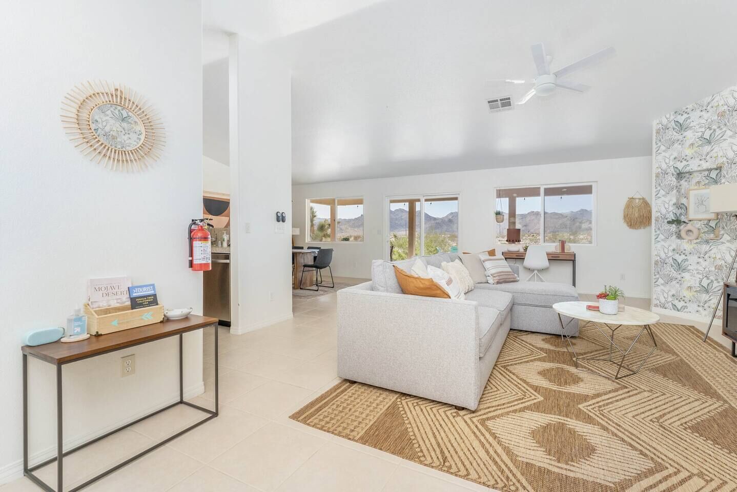 A spacious living area with a gray sofa, a small round table, and a patterned rug. A kitchen area is visible in the background with a view of the mountains.