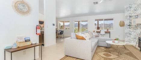A spacious living area with a gray sofa, a small round table, and a patterned rug. A kitchen area is visible in the background with a view of the mountains.