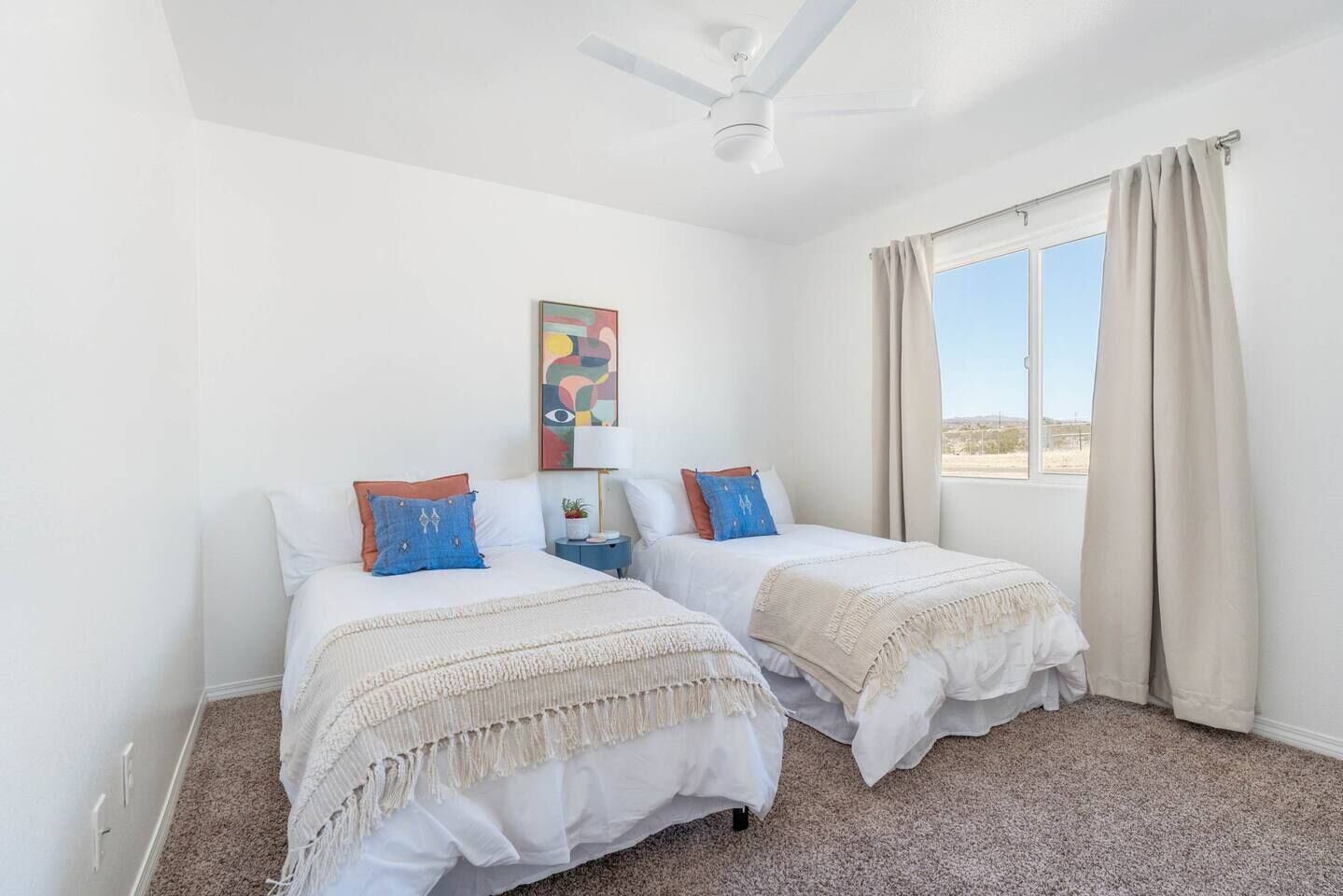A bedroom with two beds, a ceiling fan, a window with curtains, and a wall-mounted artwork.