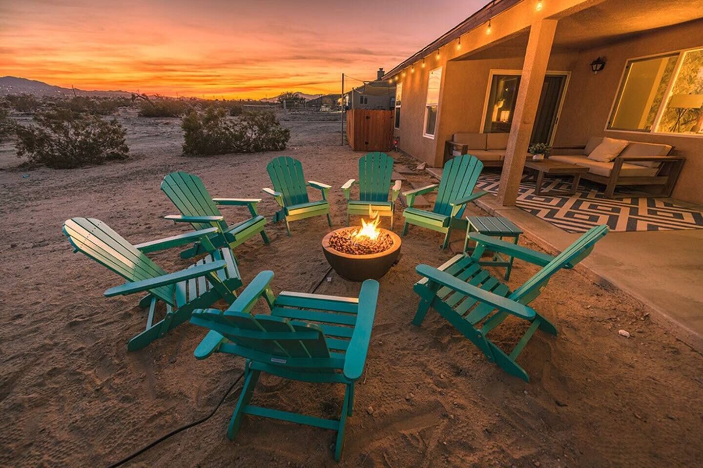 A desert landscape with a fire pit surrounded by green chairs, a covered patio area, and a building with a window overlooking the scene.