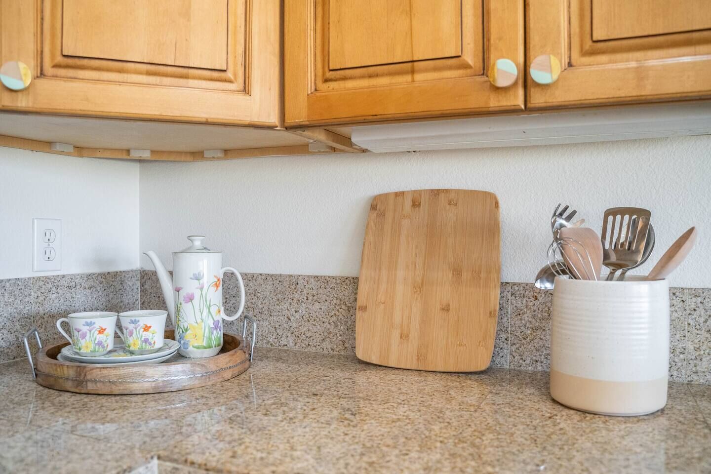 Une cuisine avec des armoires en bois, un comptoir en granit, une planche à découper en bois, une tasse blanche ornée de motifs floraux et un porte-ustensiles.