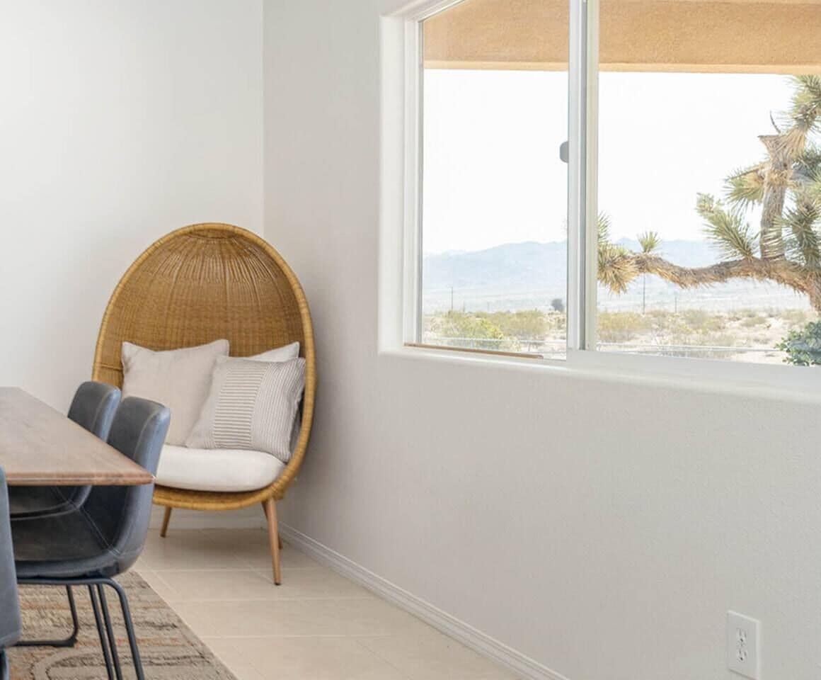 A room with a large window, a wicker chair, a table, and a view of a desert landscape.