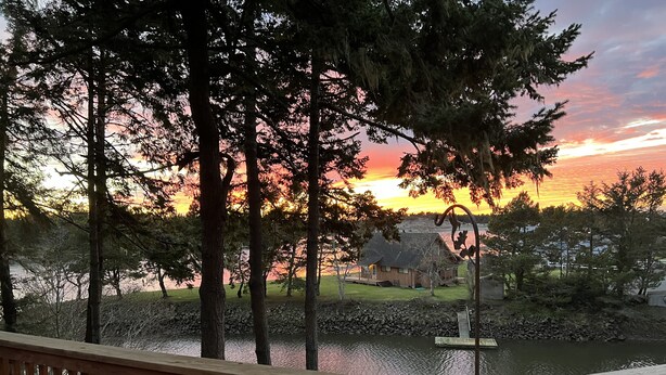 A wooden deck overlooking a lake with a sunset view, surrounded by trees and a small house. - Very quiet Pacific City home with amazing river views from the deck! (Cloverdale)