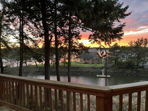 Una terraza de madera con vista a un lago al atardecer, rodeada de árboles y una pequeña casa.