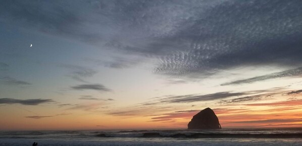 Escena de playa con una gran formación rocosa silueteada contra un cielo al atardecer.