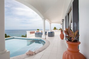 Un espace piscine avec vue sur l’océan, des colonnes blanches et des sièges d’extérieur.