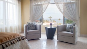 Ein Zimmer mit zwei Sesseln, einem kleinen Tisch und einem Bett. Ein großes Fenster mit transparenten Vorhängen bietet Blick auf den Strand und Palmen.