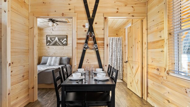 A wooden cabin interior with a dining table set for four, a bed with a mountain painting, and skis hanging on the wall. - Linville Gorge 2 Bd 1 Bth Mountain Getaway! (Newland)
