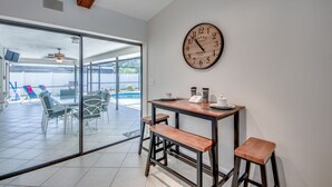 Une cuisine moderne avec une horloge en bois, une table à manger avec des tabourets et une vue sur une piscine, visible à travers des portes vitrées.