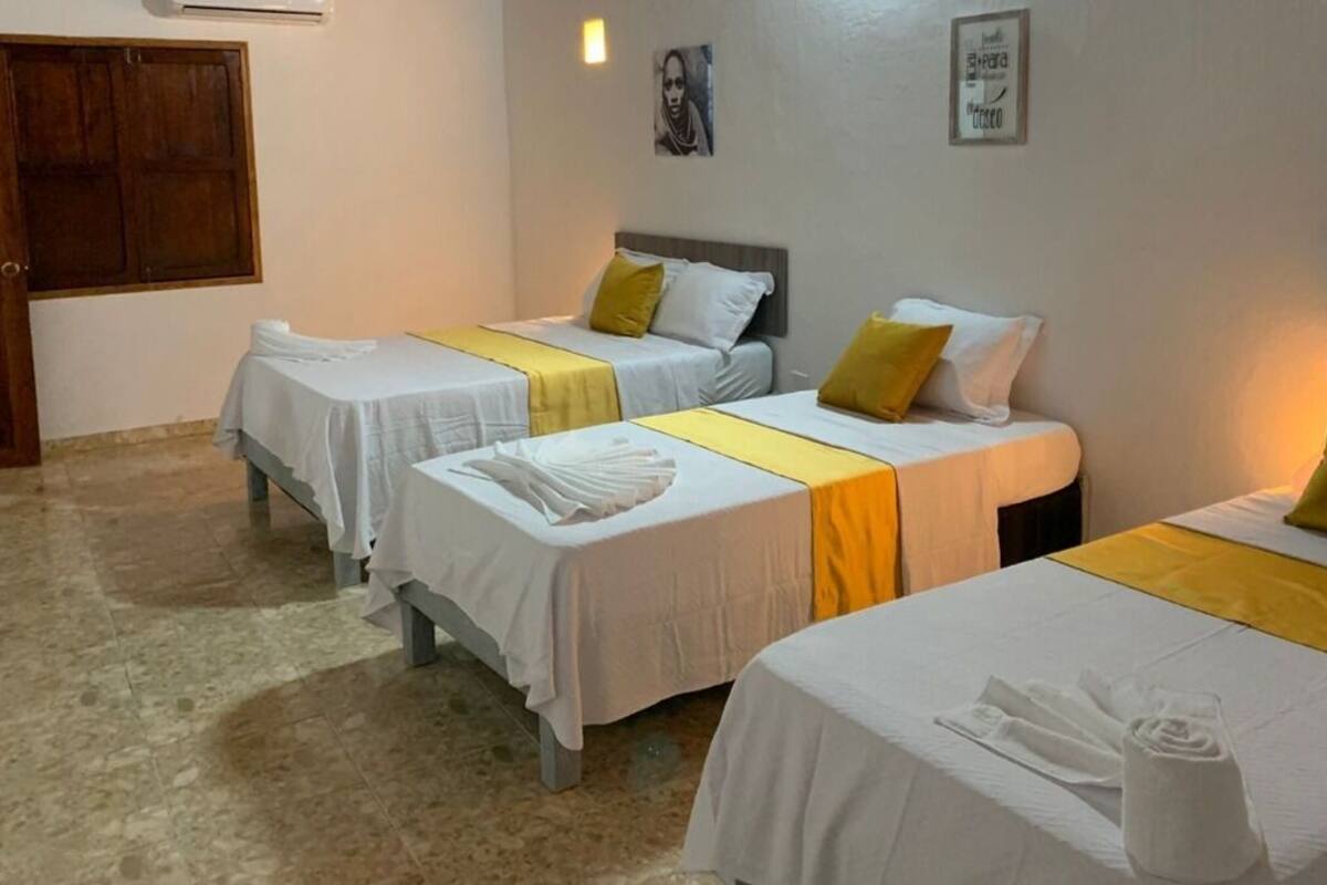 A hotel room with three single beds, each with a white bedspread and a yellow accent. There are towels on each bed, and the room has a tiled floor.