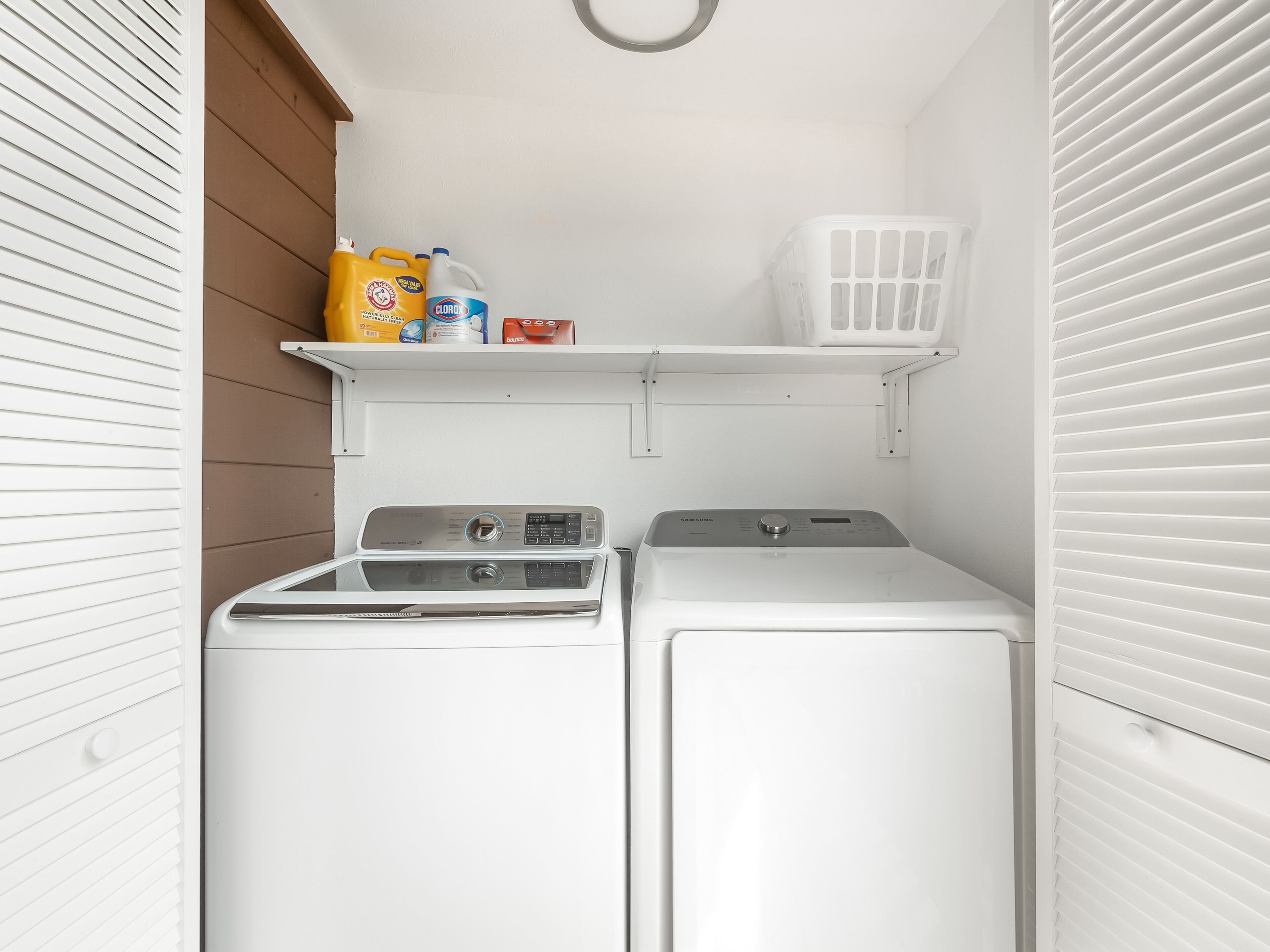 Une buanderie avec deux machines à laver blanches, une étagère avec des bouteilles de lessive et un panier à linge blanc.