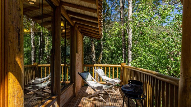 Una terraza de madera con dos sillones de descanso y una mesa, rodeada por una barandilla de madera, con vista a una zona boscosa.