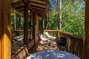Una terraza de madera con dos sillones de descanso y una mesa, rodeada por una barandilla de madera, con vista a una zona boscosa.