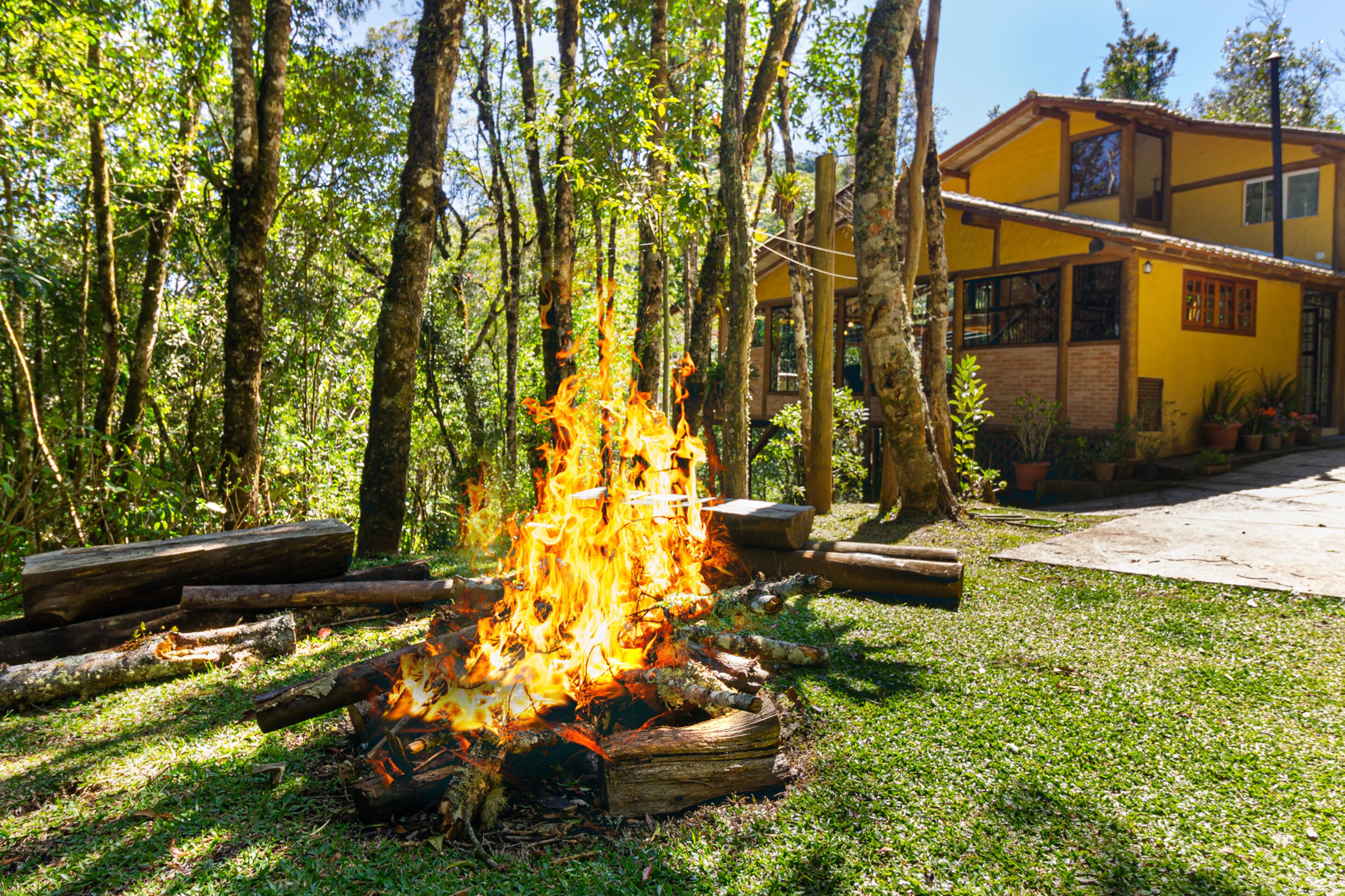 A campfire in a grassy area with trees and a yellow building in the background.