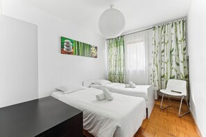 A hotel room with two beds, a desk, a chair, and a large window with bamboo curtains.