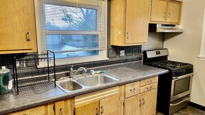 A kitchen with wooden cabinets, a stainless steel sink, a stove, and a window with blinds.