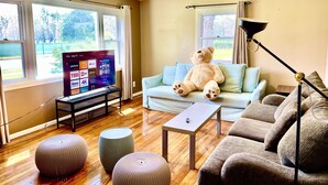 A living room with a large window, a ceiling fan, a sofa, ottomans, and a TV displaying various channels.
