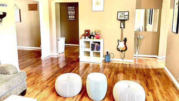 A living room with wooden flooring, a sofa, ottomans, a bookshelf, a guitar, and a teddy bear.