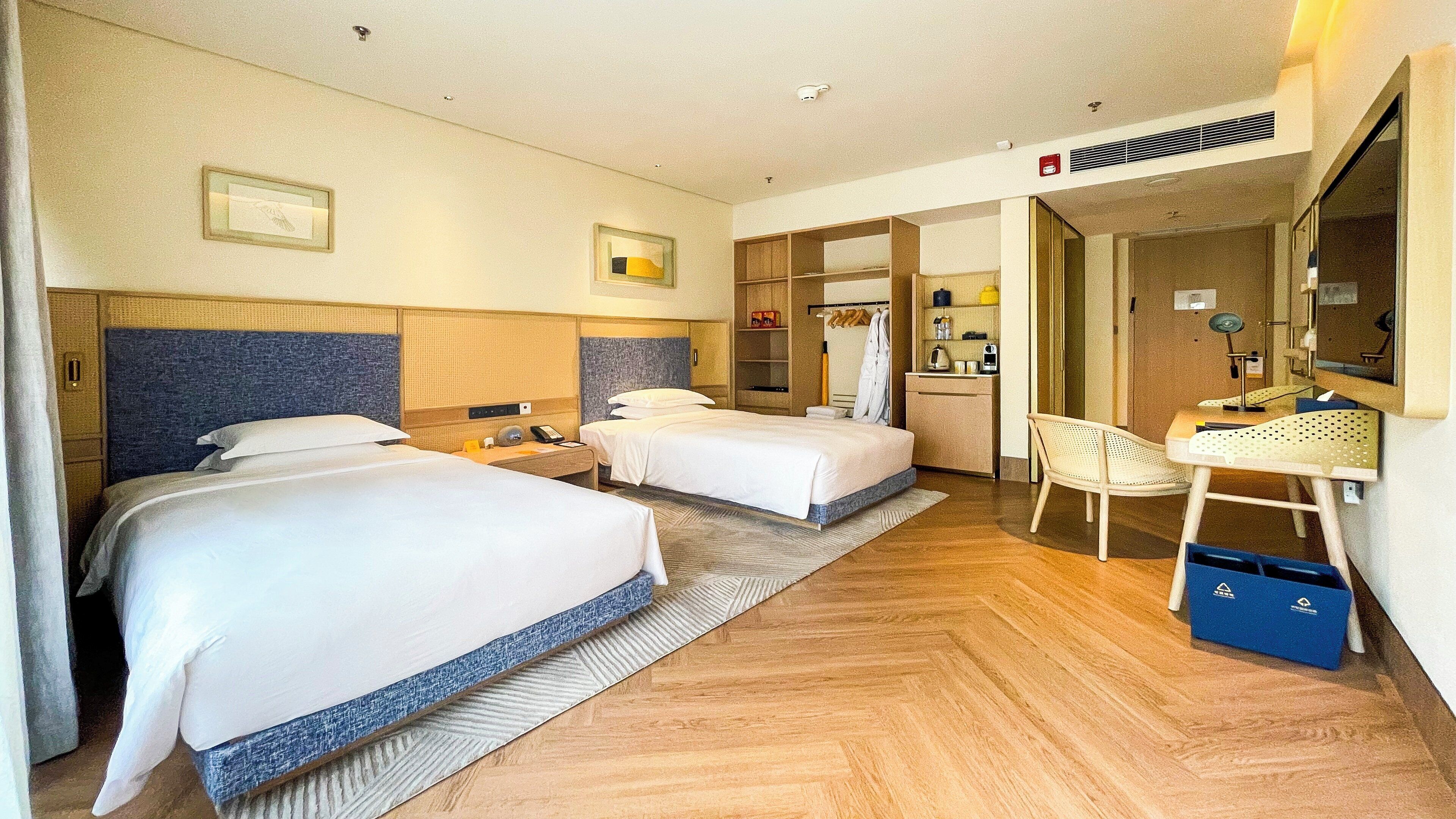 A hotel room with two beds, a desk, a chair, a mirror, and a kitchenette.
