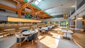 A spacious restaurant with high ceilings, wooden beams, and large windows allowing natural light. Tables and chairs are arranged for dining.