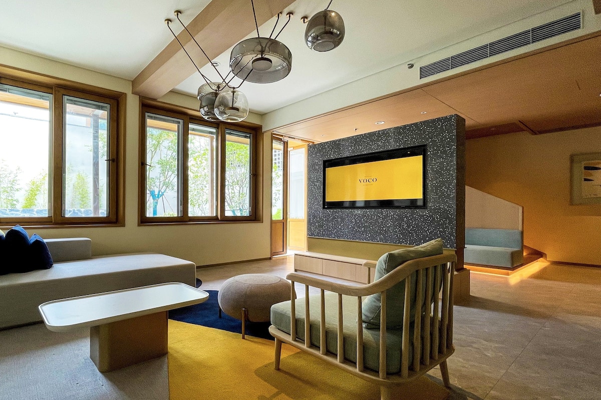 Un salon moderne avec un canapé, une table basse, un téléviseur fixé sur un mur à texture, et de grandes fenêtres.