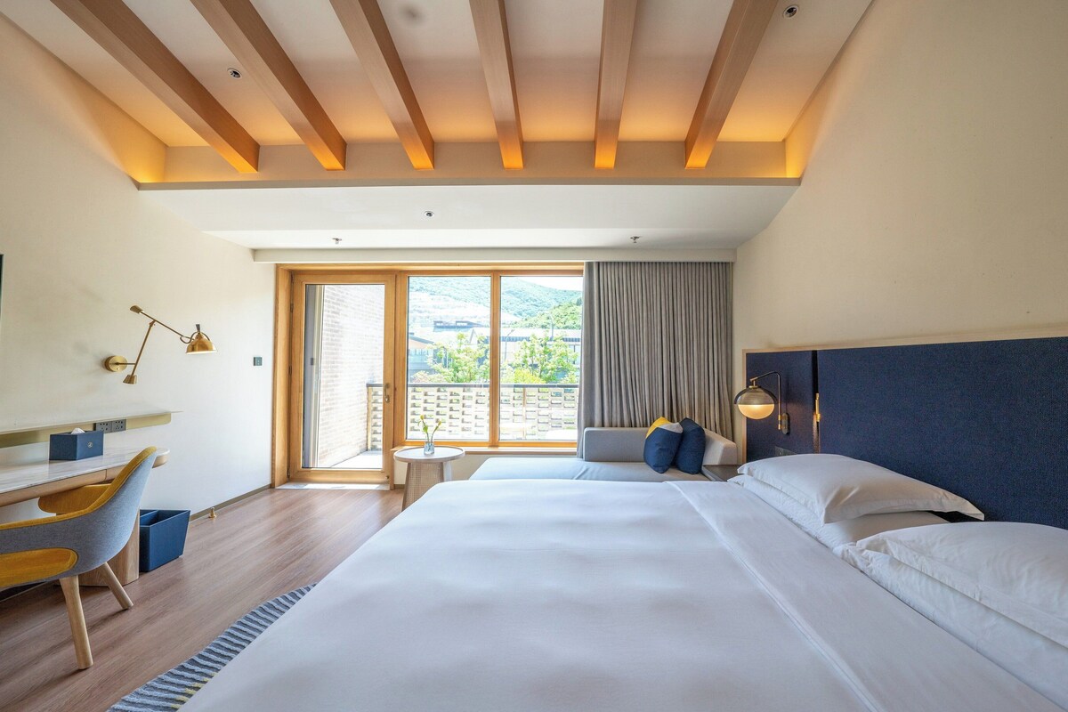 Une chambre d’hôtel avec un grand lit, un bureau avec une chaise, une télévision et une vue sur la verdure environnante.