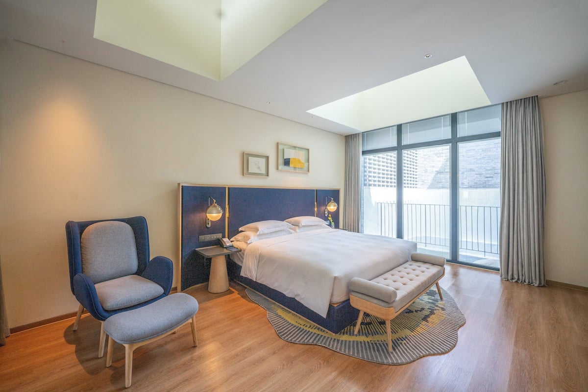 Une chambre d’hôtel moderne dotée d’un grand lit, d’un coin salon, d’un fauteuil et offrant une vue sur la ville grâce à de grandes fenêtres.