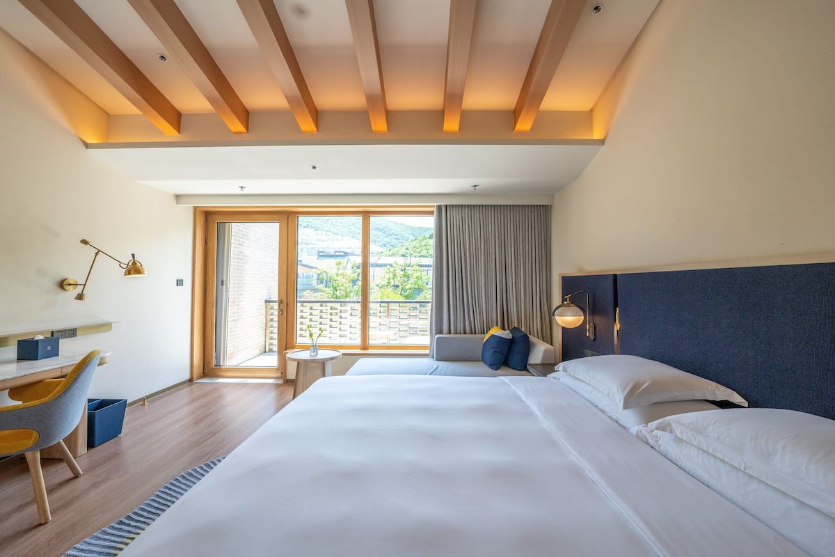 Une chambre d’hôtel avec un grand lit, un bureau avec une chaise, une télévision et une vue sur la verdure environnante.
