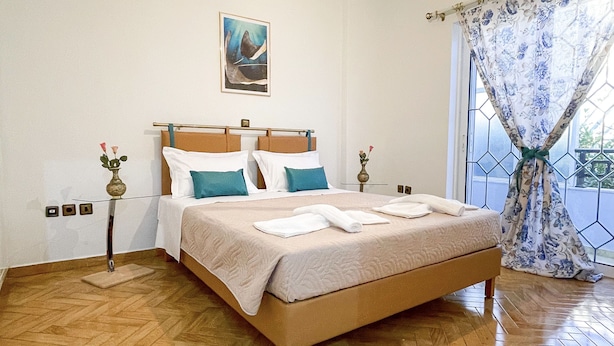 A bedroom with a wooden floor, a bed with white and teal bedding, a painting on the wall, and a window with a floral curtain.