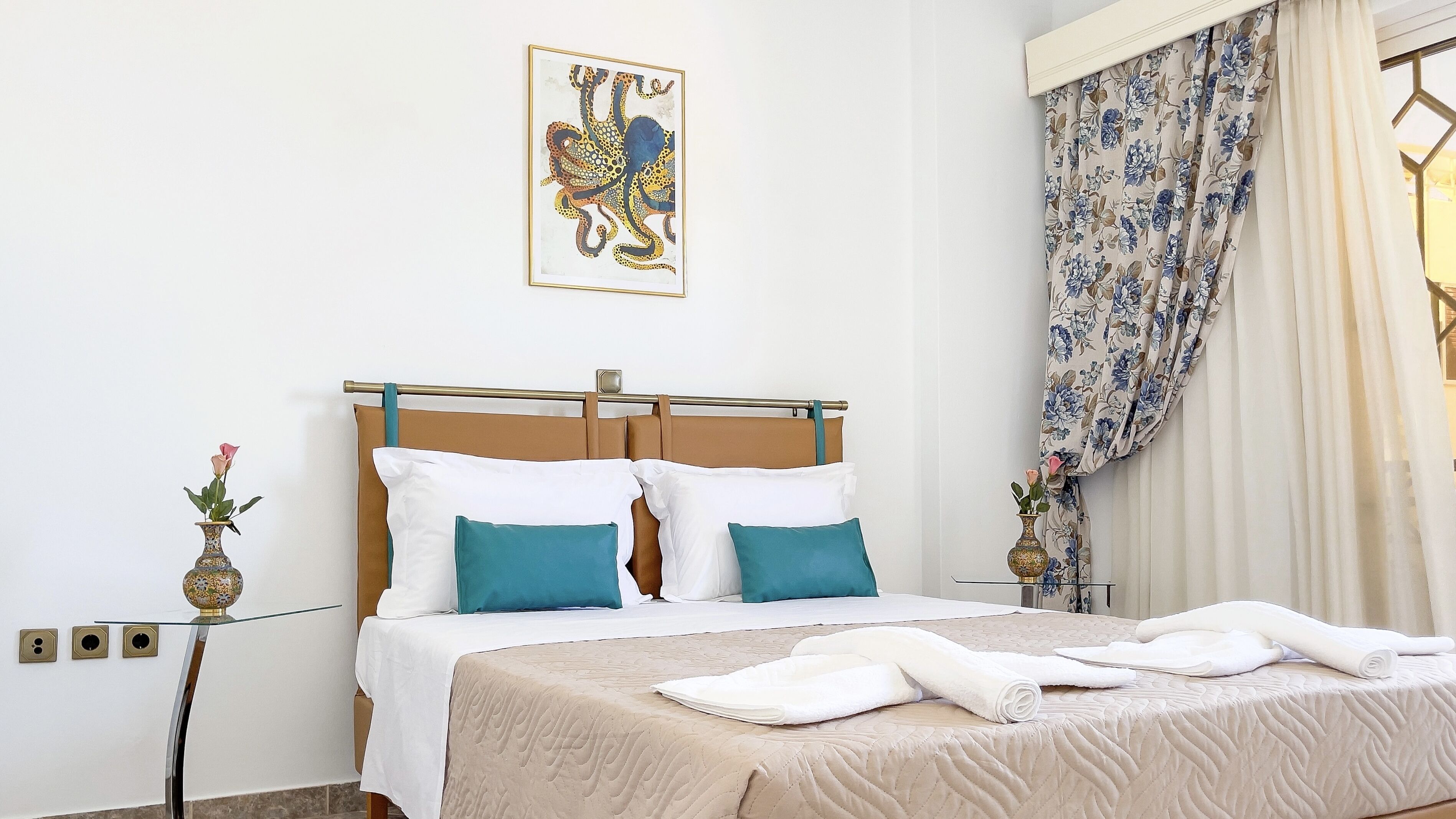 Un letto rifatto con cuscini bianchi e color acquamarina, una coperta beige e asciugamani bianchi su un tavolino in vetro.