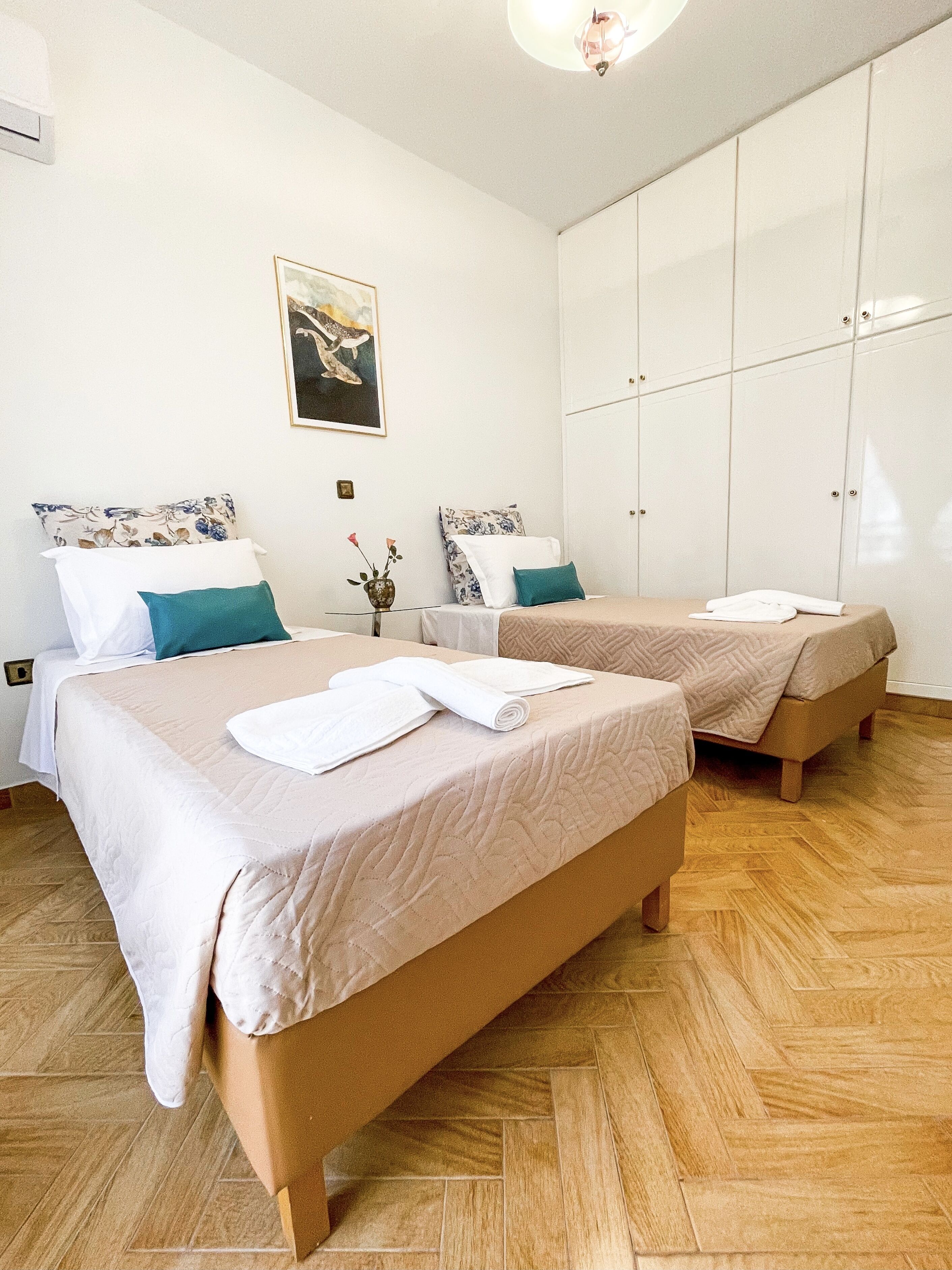 Een hotelkamer met twee bedden, elk voorzien van een wit en blauw kussen, een kleine tafel met een bloevas, en een schilderij aan de muur.