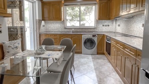 A modern kitchen with wooden cabinets, a dining table with chairs, a washing machine, and a window with a view.