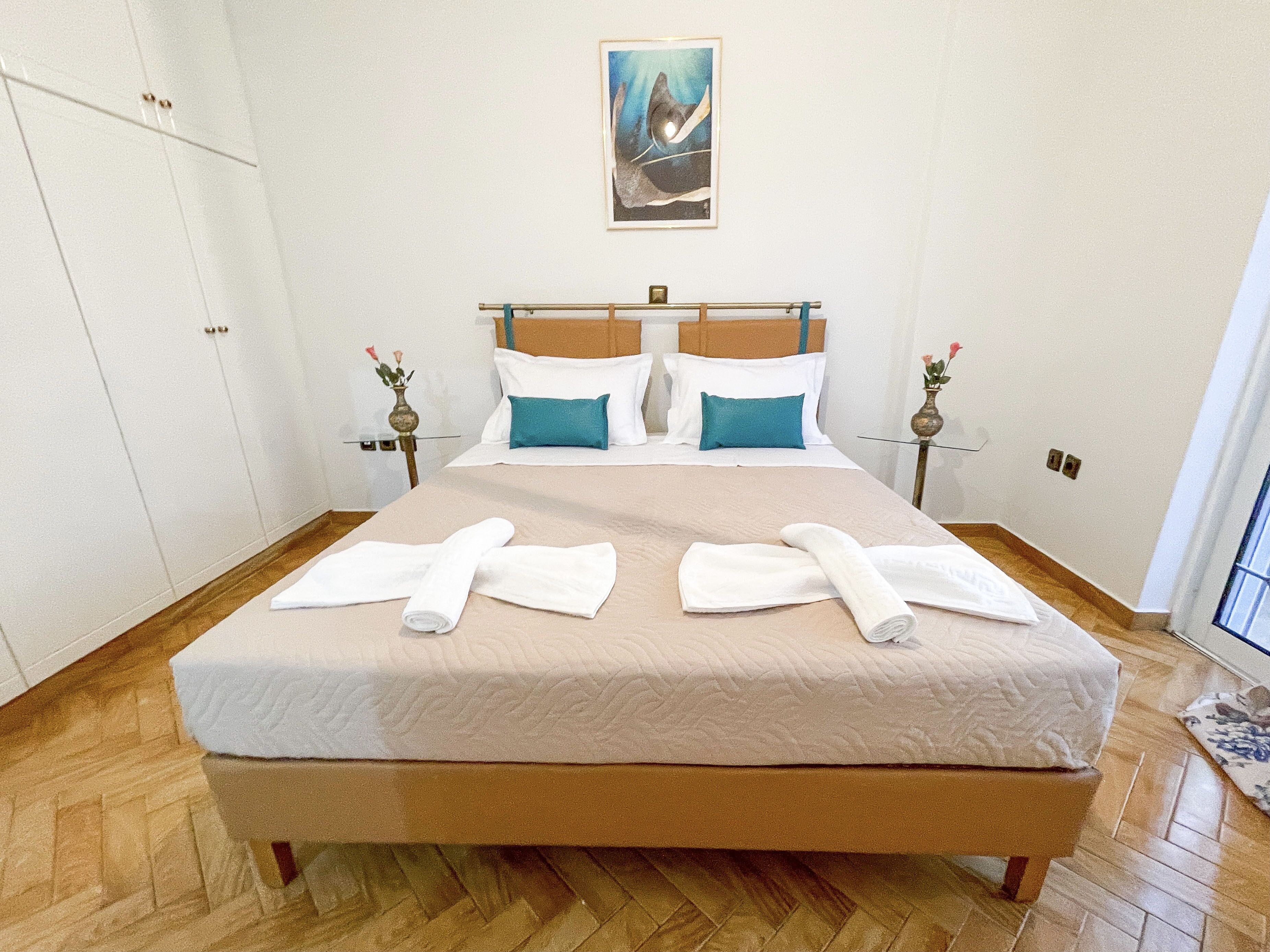A neatly made bed with white linens and two rolled towels, a wooden headboard, and a painting above the bed.