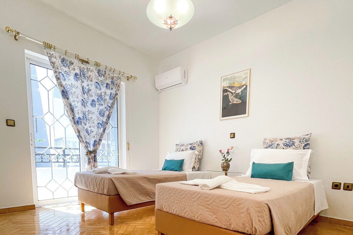 Une chambre d’hôtel avec deux lits, un parquet en bois disposé en chevrons, un ventilateur de plafond et une fenêtre ornée d’un motif géométrique.